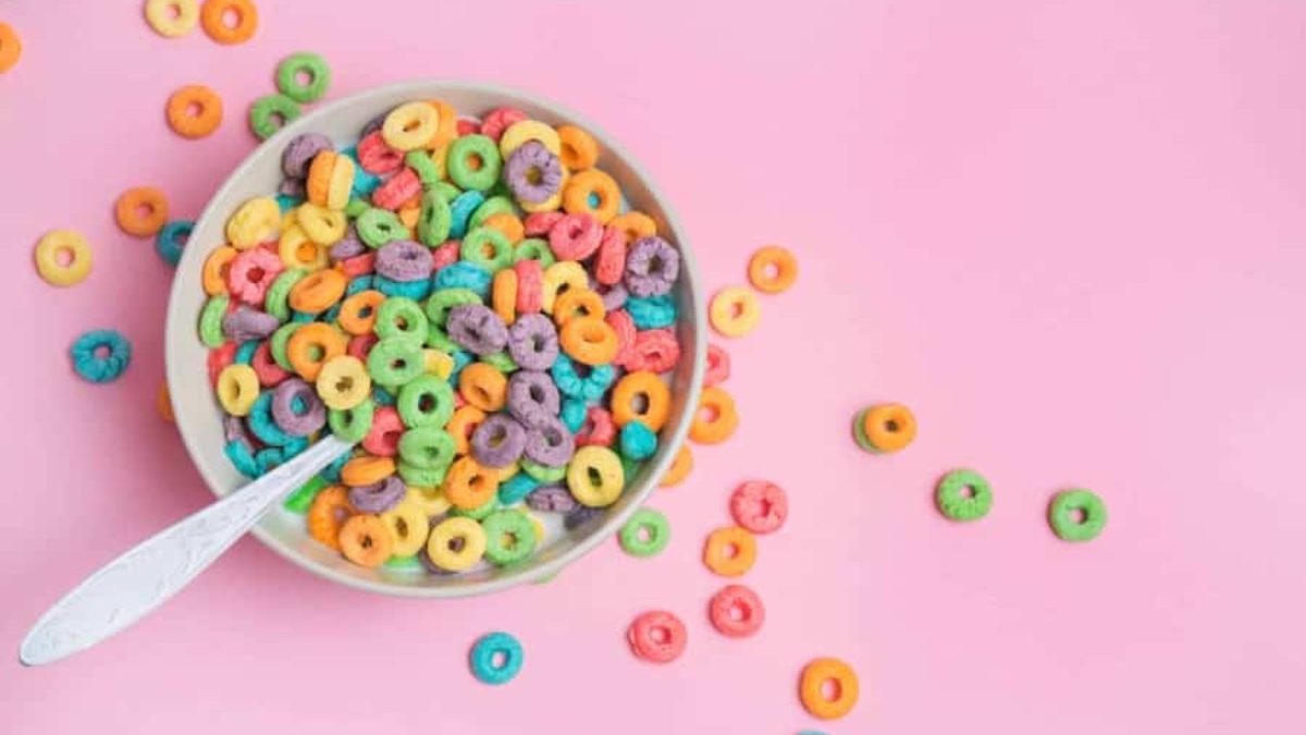 A bowl of colorful, round fruit-flavored cereal with a spoon sits on a pink background, with several cereal pieces scattered around the bowl.