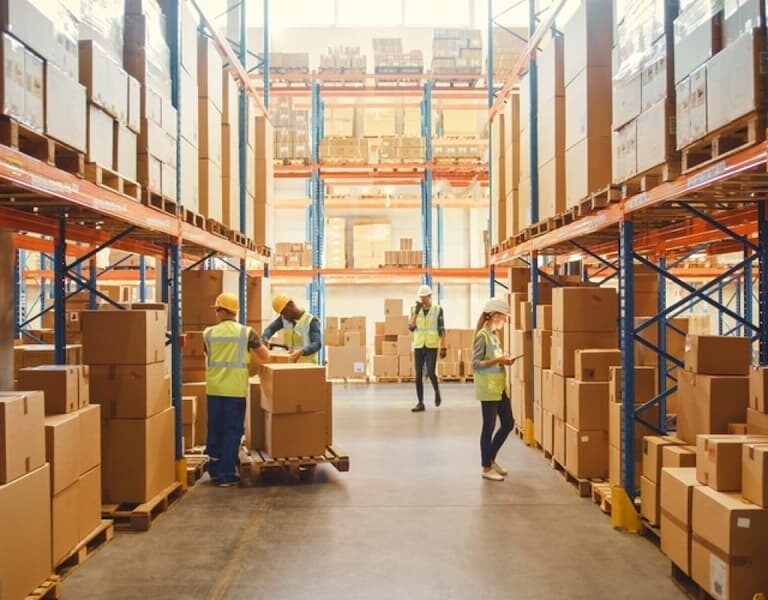 Workers wearing safety vests organize and move cardboard boxes in a large, well-lit warehouse filled with shelves stacked with more boxes.