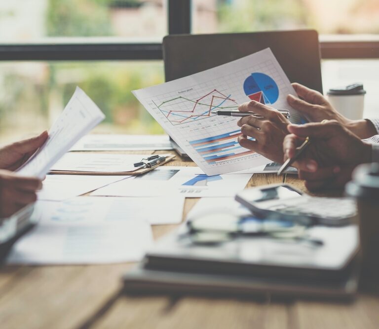 Three people sit at a table reviewing documents and charts with graphs and data. Laptops, coffee cups, and office supplies are on the table, suggesting a business meeting or team discussion in a bright office setting.