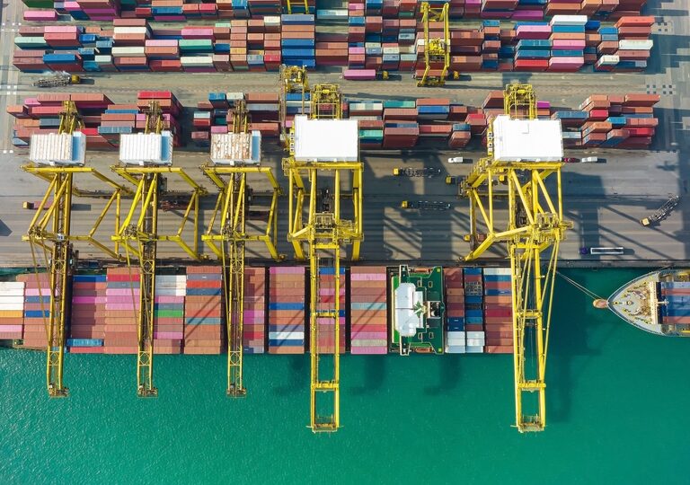 Aerial view of a busy shipping port with yellow cranes loading and unloading colorful cargo containers onto docked container ships along a turquoise waterfront.