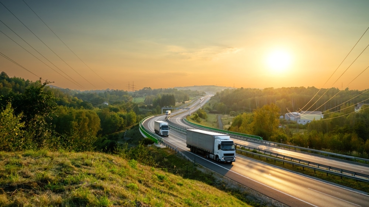 Two trucks drive on a curving highway surrounded by green trees and fields at sunrise, illustrating the efficiency outlined in a strong transportation management system business case, with power lines running alongside and a clear sky in the background.