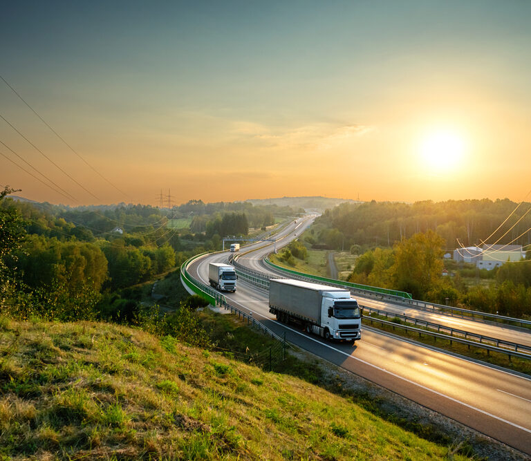 Two trucks drive on a curving highway surrounded by green trees and fields at sunrise, illustrating the efficiency outlined in a strong transportation management system business case, with power lines running alongside and a clear sky in the background.
