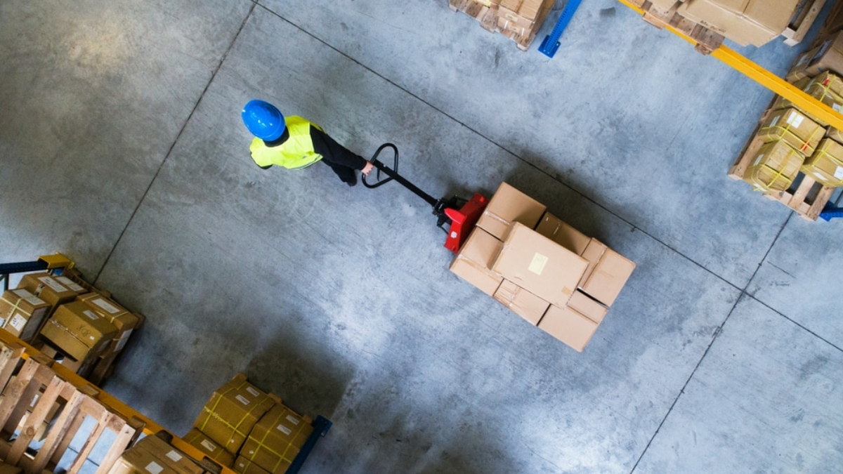 A warehouse worker wearing a blue helmet and yellow vest moves a pallet of boxes with a manual pallet jack, surrounded by stacks of cardboard boxes on pallets, viewed from above.