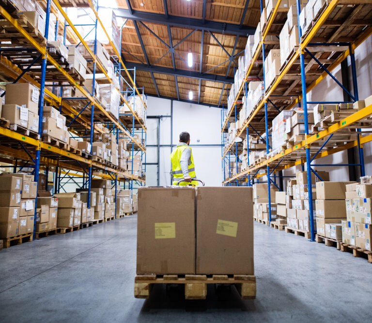 A worker in a high-visibility vest stands in a large ab701 warehouse filled with stacked boxes on shelves, with two large boxes on a pallet in the foreground.