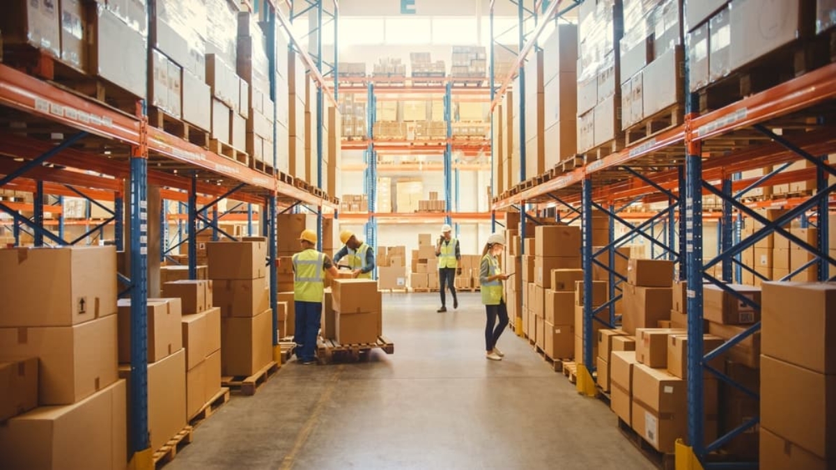 Warehouse workers in safety vests organize and move large cardboard boxes along wide aisles lined with tall metal shelves, efficiently tracking inventory with a WMS. Bright lighting and high ceilings are visible.