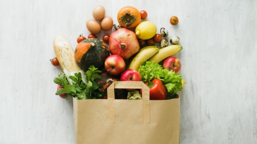 A brown paper bag overflowing with fresh groceries, including eggs, bread, pumpkins, apples, bananas, lettuce, tomatoes, a pomegranate, lemons, strawberries, mint, and quail eggs on a white surface—perfect for FSMA-compliant kitchens.