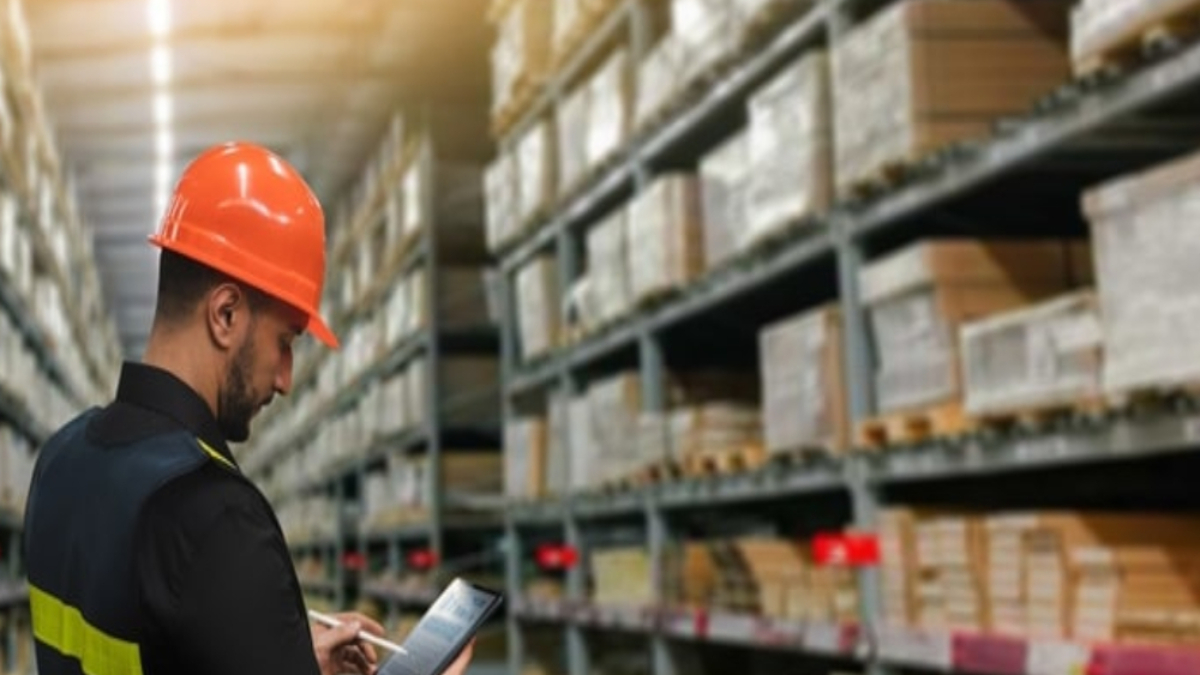 A worker wearing an orange hard hat and safety vest uses a tablet in a warehouse filled with shelves stacked with boxes and packages.