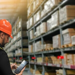 A worker wearing an orange hard hat and safety vest uses a tablet in a warehouse filled with shelves stacked with boxes and packages.