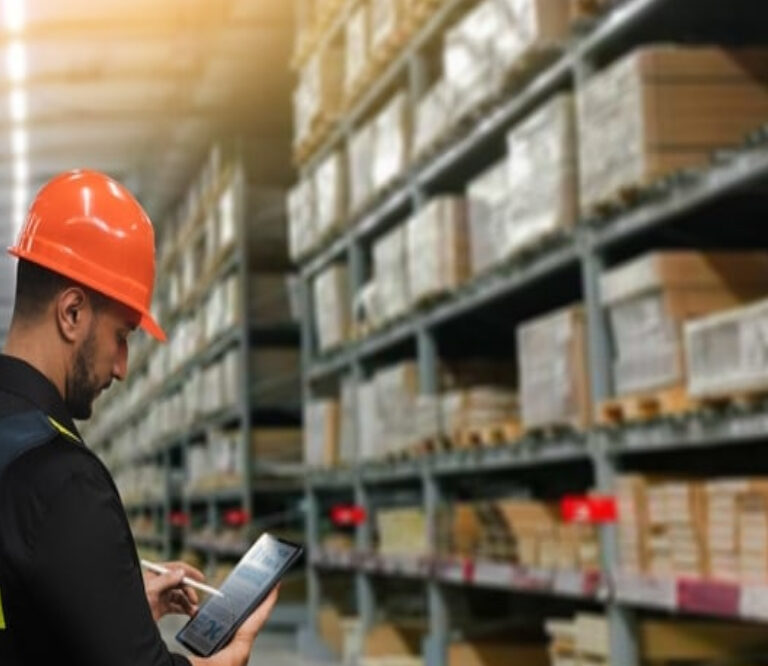 A worker wearing an orange hard hat and safety vest uses a tablet in a warehouse filled with shelves stacked with boxes and packages.