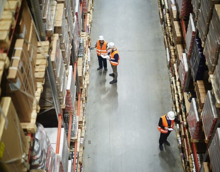 Overhead view of three workers in orange safety vests and hard hats walking and inspecting shelves in a large warehouse filled with stacked boxes and packages.