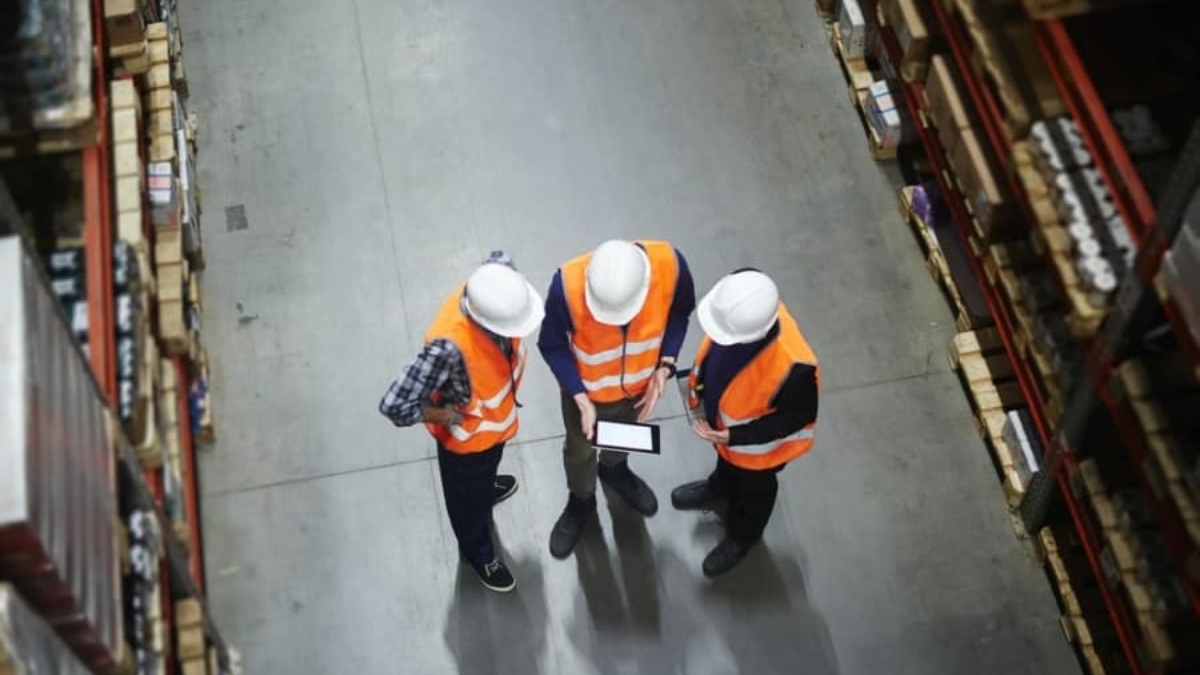 Three people wearing white hard hats and orange safety vests stand in a warehouse aisle, looking at a tablet. Shelves stocked with boxes and items are visible on both sides.