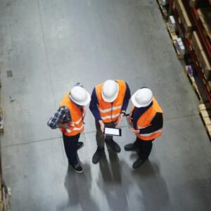 Three people wearing white hard hats and orange safety vests stand in a warehouse aisle, looking at a tablet. Shelves stocked with boxes and items are visible on both sides.