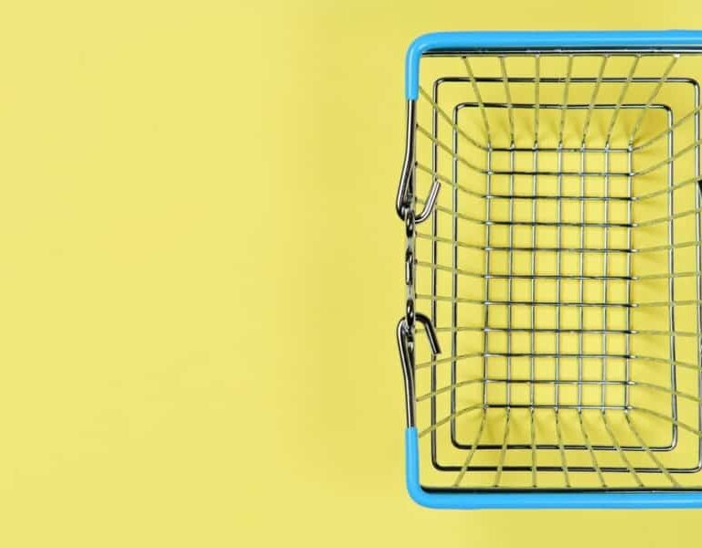 A small, empty metal shopping basket with blue handles sits on a yellow background, viewed from above.