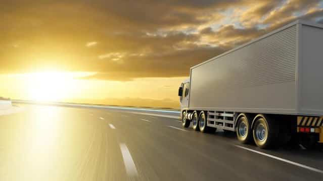 A large white semi-truck drives down a highway at sunset, with the golden sun low on the horizon and the sky filled with dramatic clouds. The road ahead is empty.