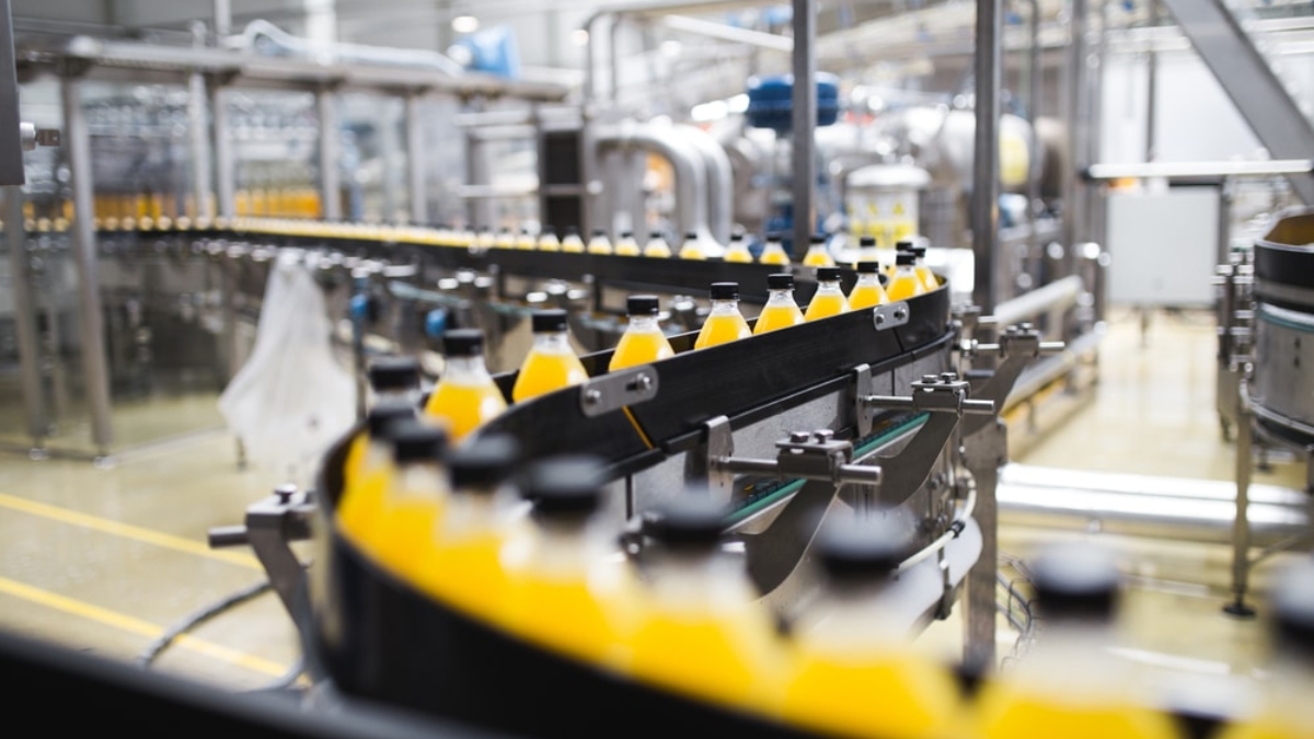 A conveyor belt carries rows of plastic bottles filled with orange liquid through a modern beverage factory, where automation strategies and stainless steel machinery ensure efficient production.