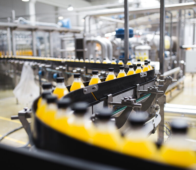 A conveyor belt carries rows of plastic bottles filled with orange liquid through a modern beverage factory, where automation strategies and stainless steel machinery ensure efficient production.