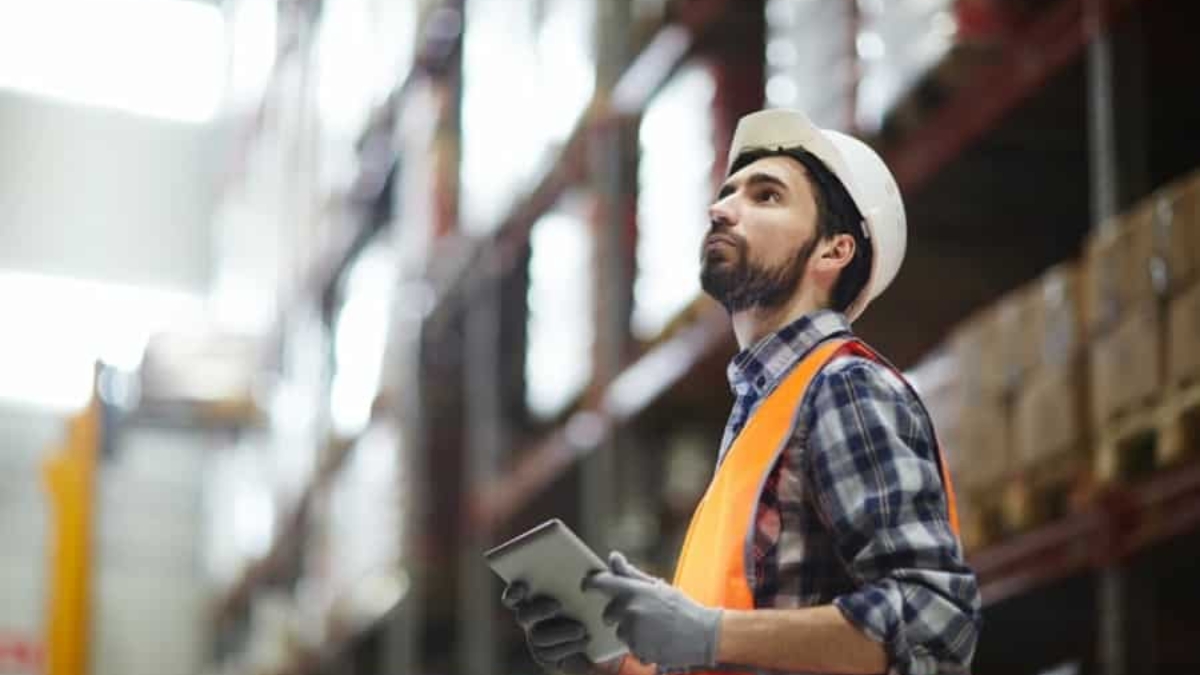 A warehouse worker in a hard hat, orange safety vest, and gloves holds a tablet, possibly checking a Warehouse Management System (WMS) Cheat Sheet while looking up at shelves full of inventory in a blurred industrial setting.