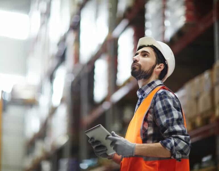 A warehouse worker in a hard hat, orange safety vest, and gloves holds a tablet, possibly checking a Warehouse Management System (WMS) Cheat Sheet while looking up at shelves full of inventory in a blurred industrial setting.