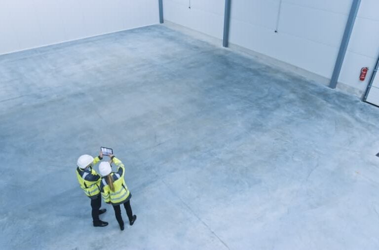 Two people in safety vests and helmets stand together in a large, empty industrial warehouse, looking at a tablet. The warehouse has concrete floors and high white walls.