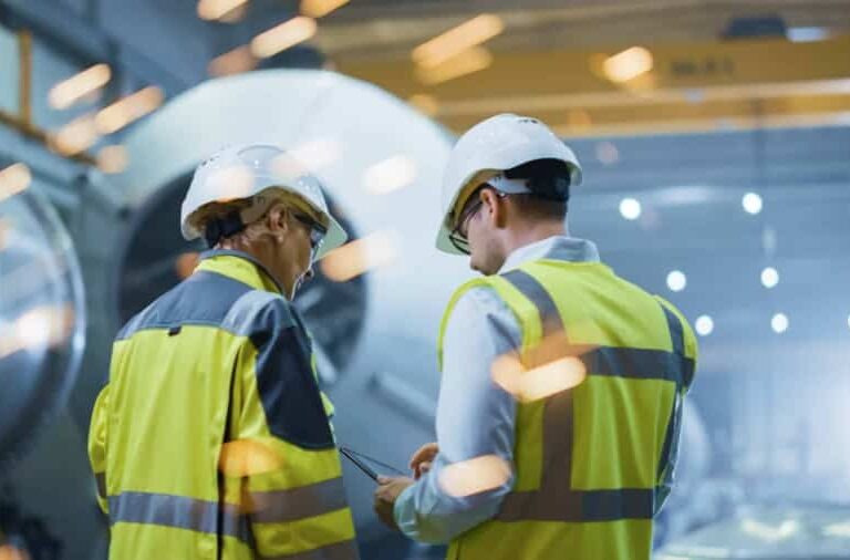 Two workers wearing safety vests and helmets stand in an industrial setting, with machinery and bright flying sparks in the background, suggesting active manufacturing or metalworking.