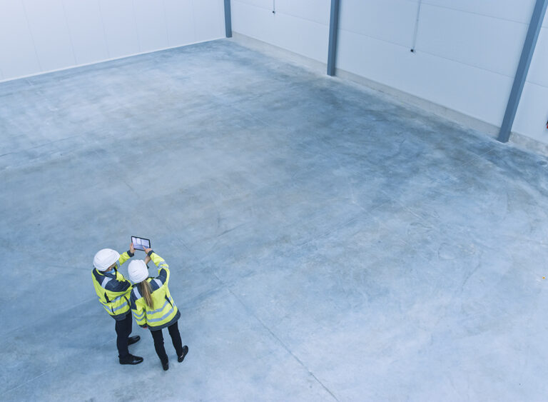 Two construction workers in high-visibility jackets and hard hats stand in an empty, spacious industrial warehouse, looking at a tablet together. The warehouse has a concrete floor and white walls.