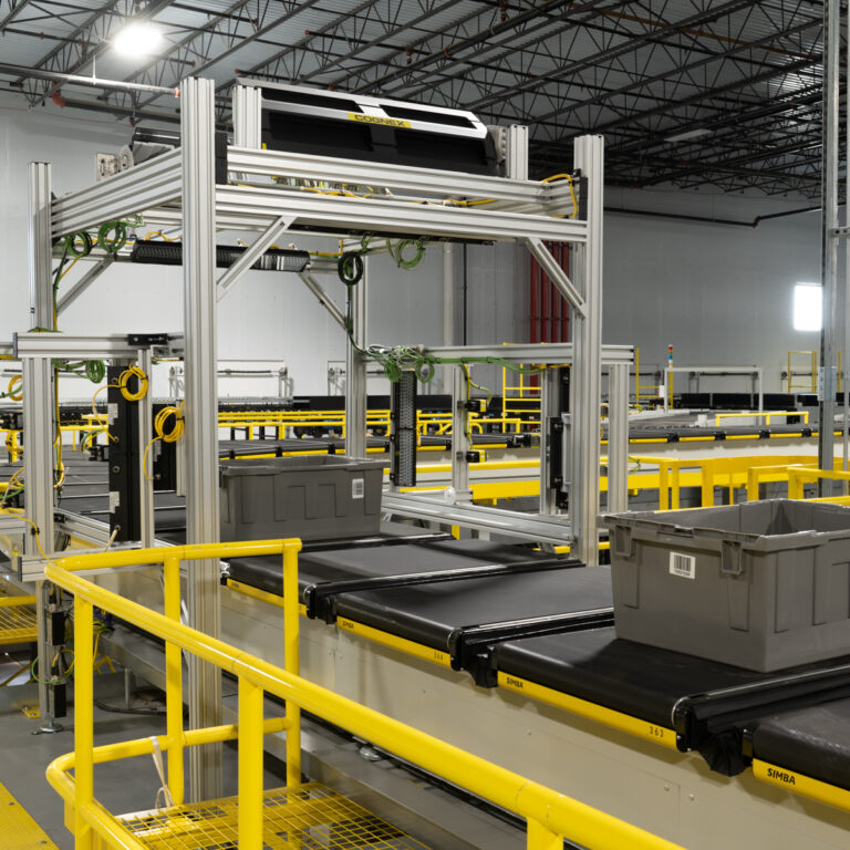 A gray plastic bin moves along a conveyor belt system inside a large industrial warehouse with yellow safety railings and exposed ceiling beams.