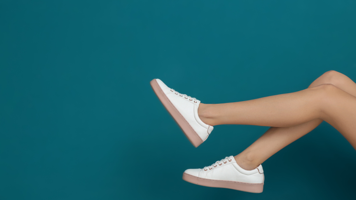 A person with crossed legs wears white sneakers with pink soles, shown against a plain teal background. The focus is on the lower legs and shoes.
