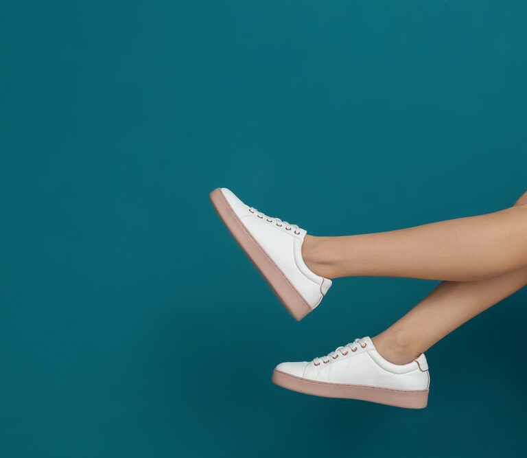 A person with crossed legs wears white sneakers with pink soles, shown against a plain teal background. The focus is on the lower legs and shoes.