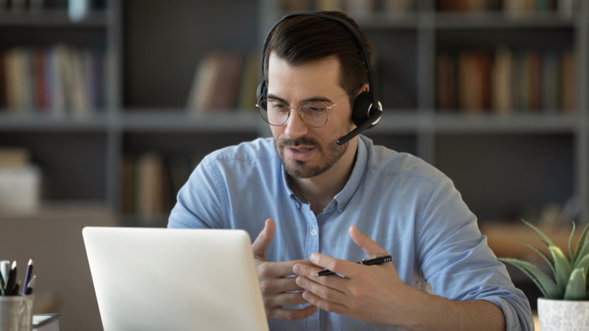 A man wearing glasses and a headset sits at a desk in front of a laptop, gesturing with his hands while speaking during a video call about Microsoft’s Sales Copilot. Bookshelves are visible in the background.