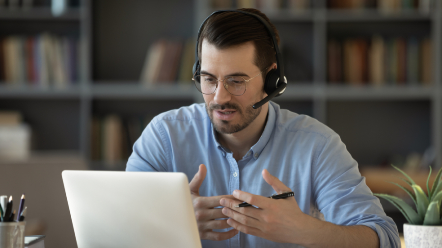 A man wearing glasses and a headset sits at a desk in front of a laptop, gesturing with his hands while speaking during a video call about Microsoft’s Sales Copilot. Bookshelves are visible in the background.
