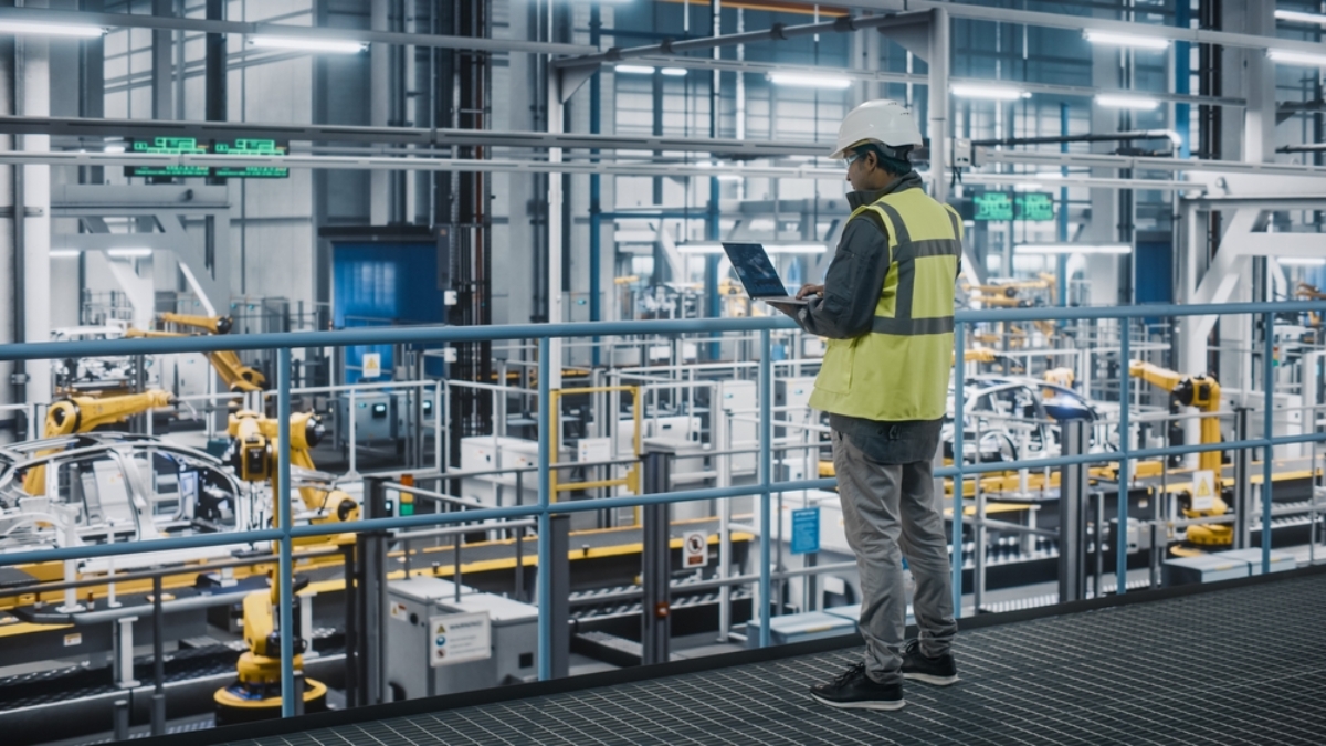 A worker in a yellow safety vest and hard hat stands on a balcony overlooking an automated factory floor, using a laptop to monitor the WMS while observing robotic arms and machinery operating below.