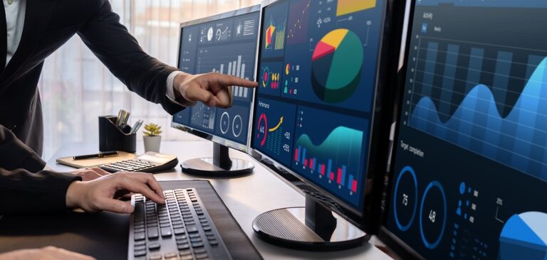 Two people analyze colorful financial charts and data visualizations on multiple monitors in an office, utilizing Microsoft Business Solutions—one points at a screen while the other types on a keyboard.