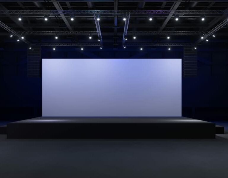 A large, empty stage with a blank screen is illuminated by spotlights in a dark auditorium, setting the scene for a WMS Implementation Case Study presentation or performance.