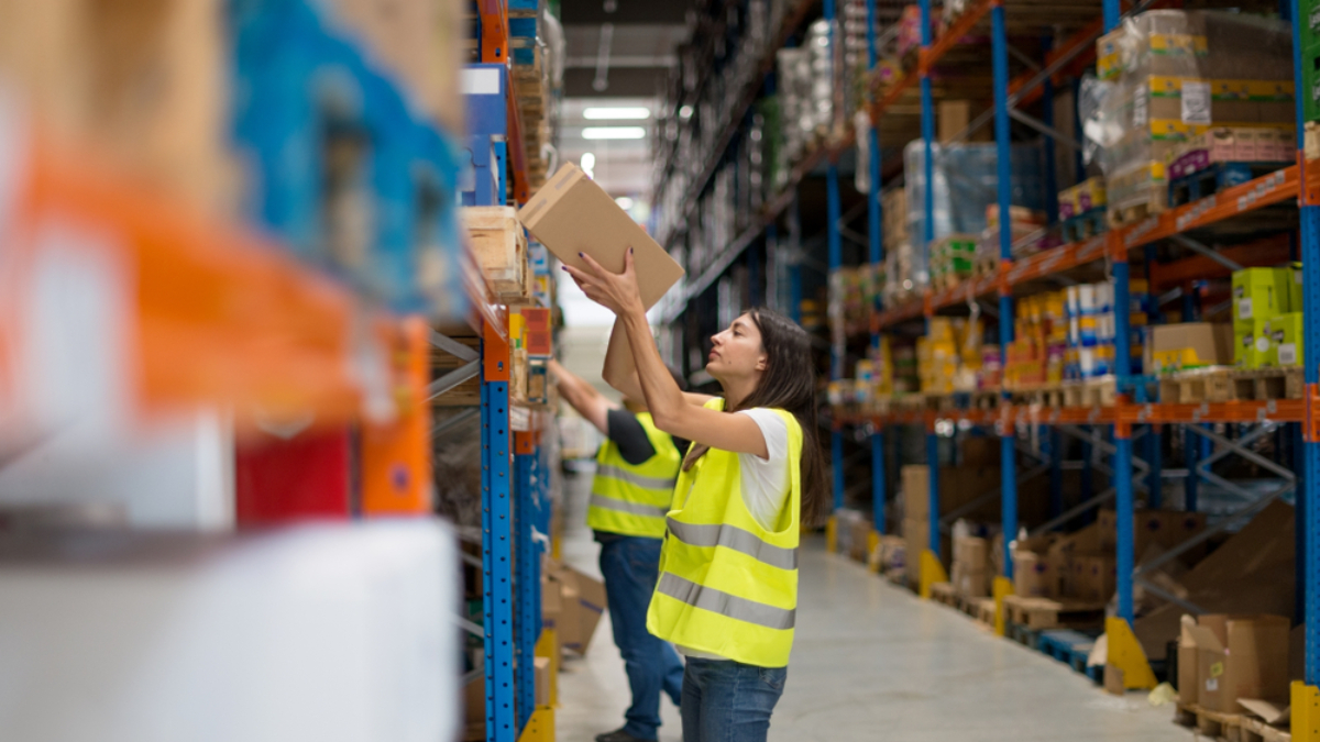 A worker in a yellow safety vest reaches up to pick a box from a shelf in a large warehouse filled with stocked shelves. Another worker is visible in the background, also organizing items.