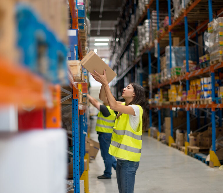A worker in a yellow safety vest reaches up to pick a box from a shelf in a large warehouse filled with stocked shelves. Another worker is visible in the background, also organizing items.