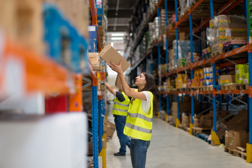 A worker in a yellow safety vest reaches up to pick a box from a shelf in a large warehouse filled with stocked shelves. Another worker is visible in the background, also organizing items.