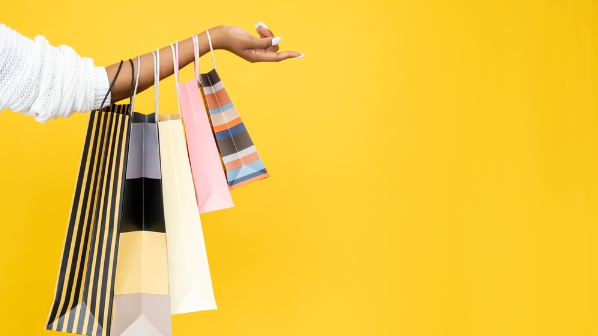A persons outstretched arm holds several colorful shopping bags against a bright yellow background. The bags have various patterns and colors, including stripes and solid tones.