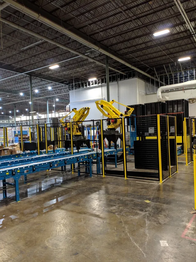 A factory interior with two yellow robotic arms behind safety fences, next to conveyor belts featuring Hytrol replacement parts on a shiny concrete floor under high ceilings with exposed beams and bright overhead lighting.