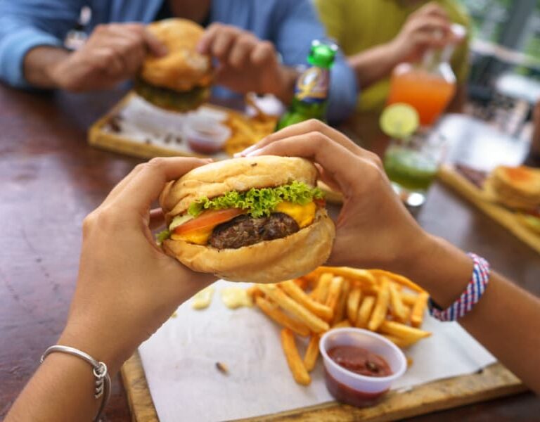 A person holds a cheeseburger with lettuce and tomato over a tray of fries and ketchup, with other people eating burgers and drinking colorful beverages at a wooden table.