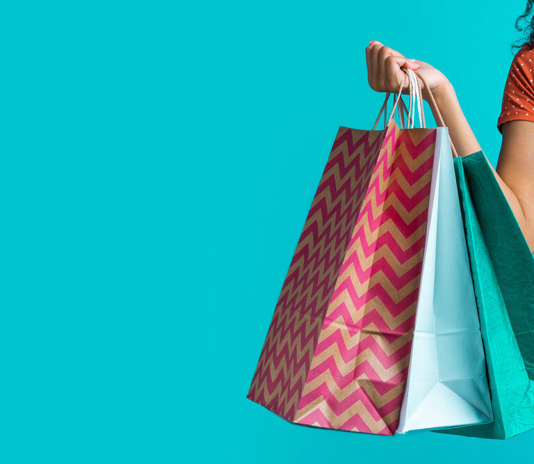 A person wearing an orange polka dot shirt holds colorful shopping bags against a bright turquoise background. Only the persons arm and part of their torso are visible.