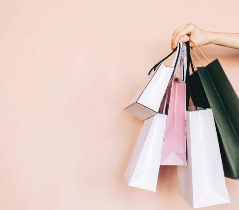 A hand is holding several colorful shopping bags by their handles against a light peach background, highlighting the variety that WMS can help organize. The bags vary in size and color, including white, pink, black, and dark green.