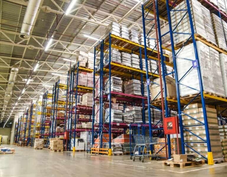 Large warehouse interior with tall blue and yellow racks stacked with pallets of boxes and goods. Wide aisles separate the shelves, and bright lighting illuminates the organized storage area.