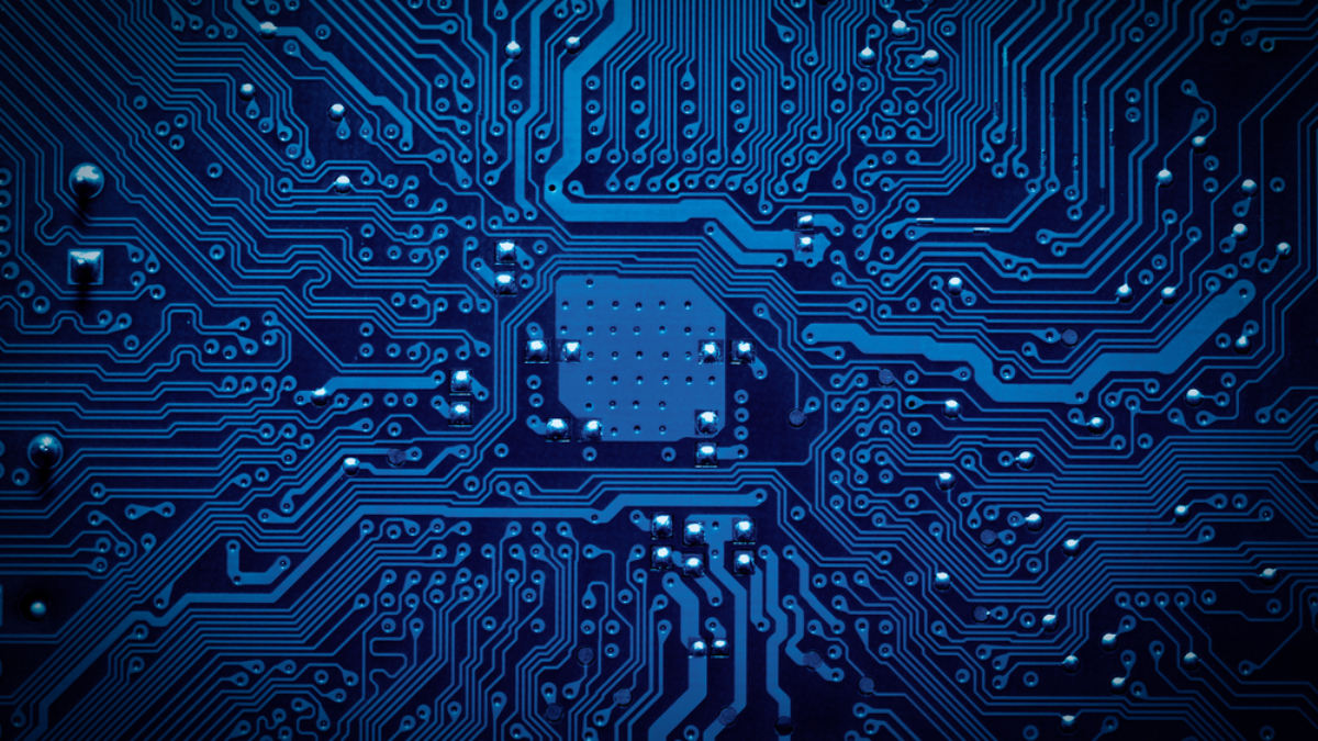 Close-up of a blue computer circuit board showing intricate electronic pathways and components, including a central microchip area with numerous connecting lines and soldering points.