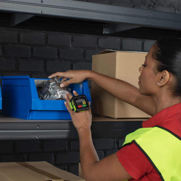 A woman wearing a reflective vest scans a metal part using a handheld barcode scanner while organizing items in blue plastic bins on a warehouse shelf. Cardboard boxes are visible nearby.