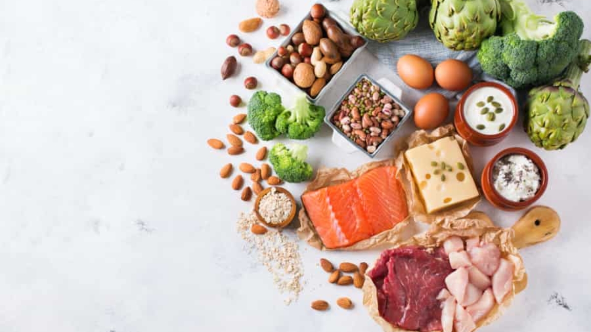 Assorted healthy foods including salmon, beef, chicken, cheese, eggs, nuts, seeds, yogurt, artichokes, broccoli, and beans arranged on a white surface.