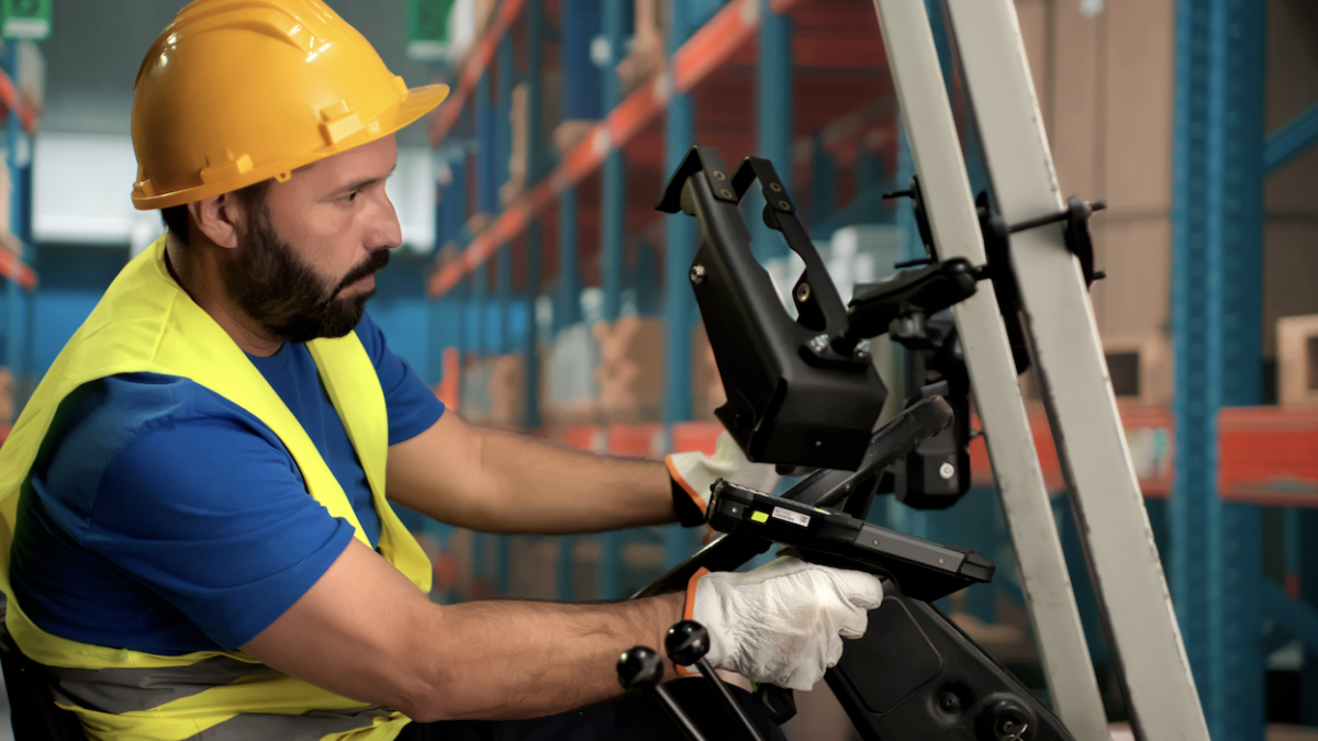 A warehouse worker wearing a yellow hard hat, safety vest, and gloves operates a forklift in a storage area with shelves filled with boxes, efficiently managing inventory using an ERP system.