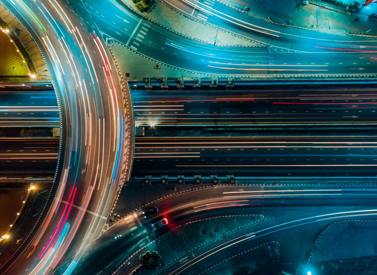 Aerial view of a brightly lit highway interchange at night, with streaks of blue and red lights from moving vehicles creating dynamic lines and patterns on the roads.