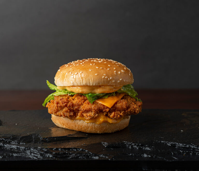 A crispy fried chicken sandwich with lettuce, cheddar cheese, and sauce on a sesame seed bun, placed on a dark slate surface with a plain dark background.