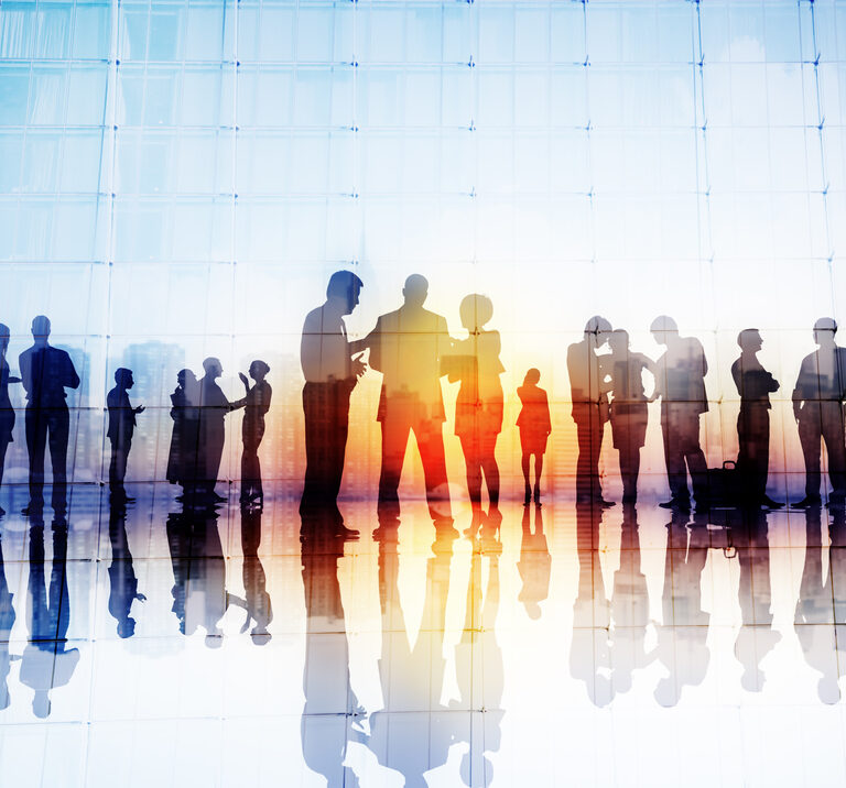 Silhouettes of business people standing and talking in groups against a large glass window, reflecting on the shiny floor as they discuss S&OP strategies in the soft sunset light.