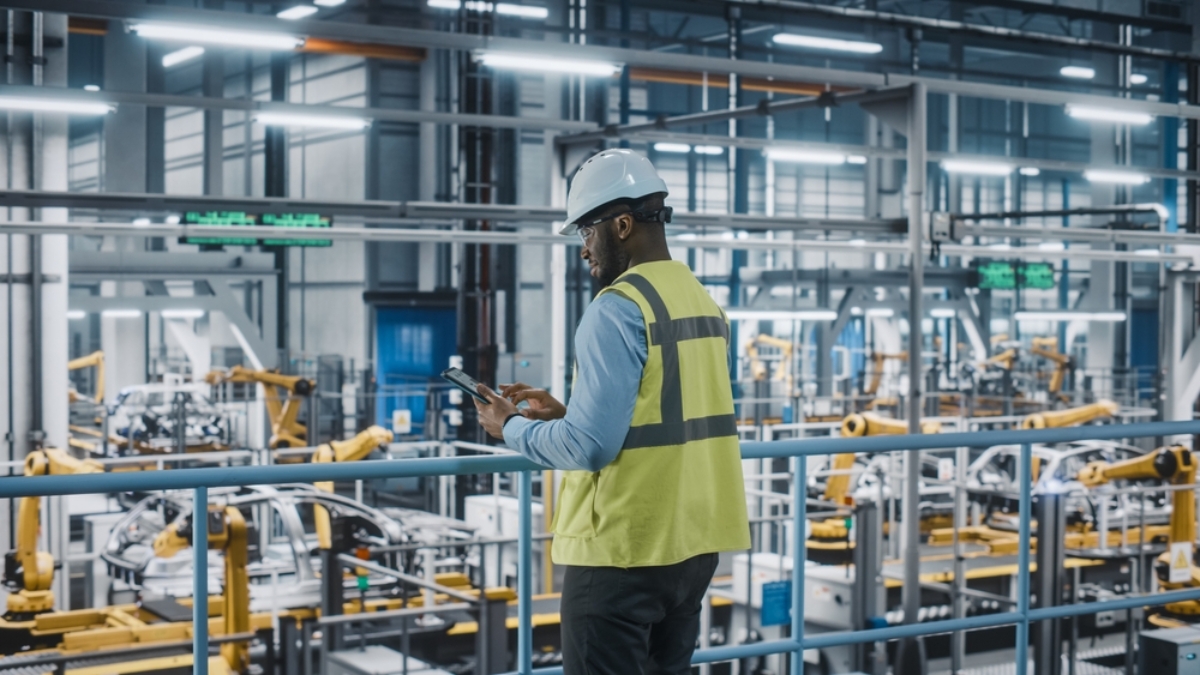 A person wearing a white hard hat and yellow safety vest uses a tablet while standing in a modern, automated factory with robotic arms and machinery in the background.